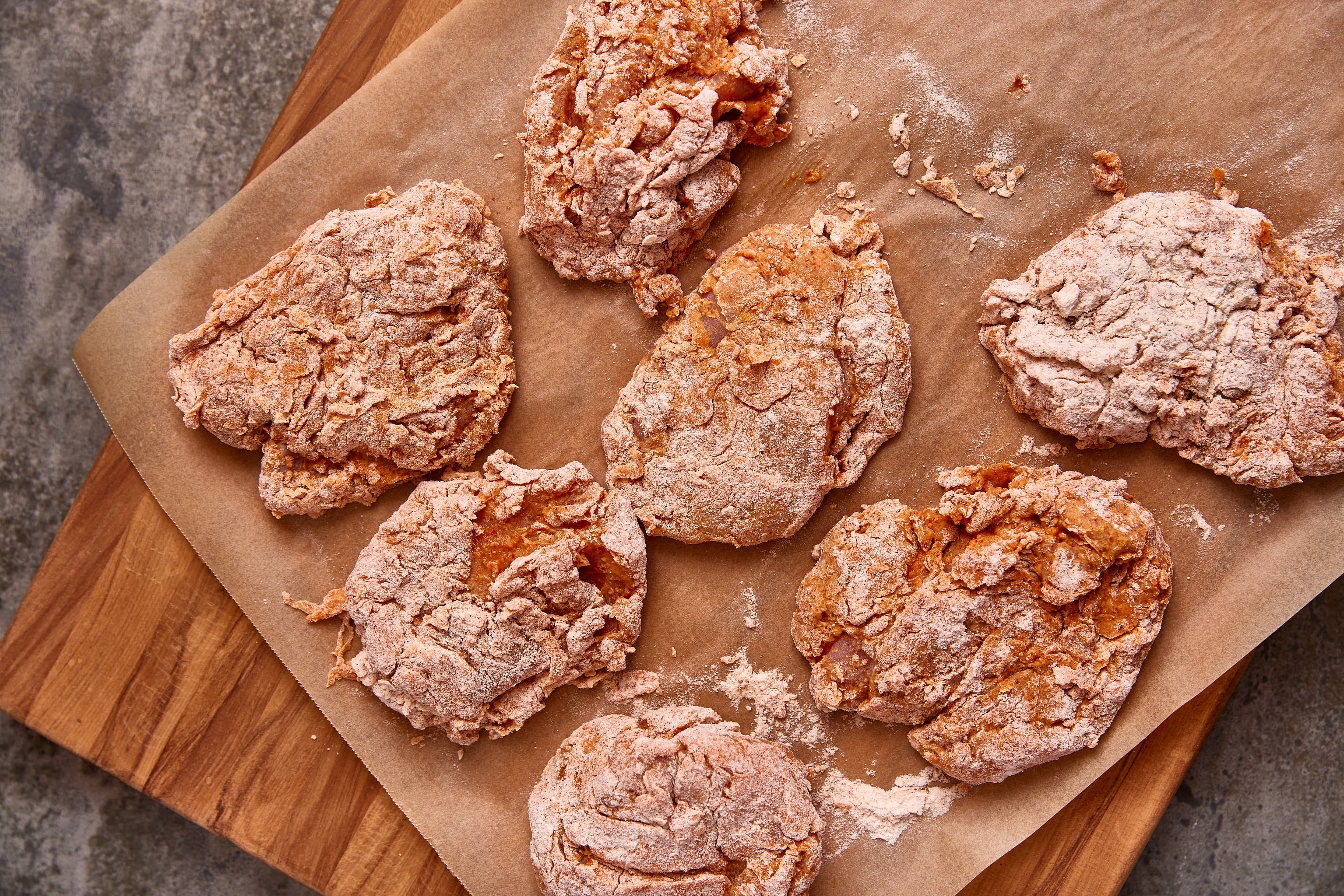 Coated chicken pieces on a wooden cutting board with brown paper underneath before being fried.