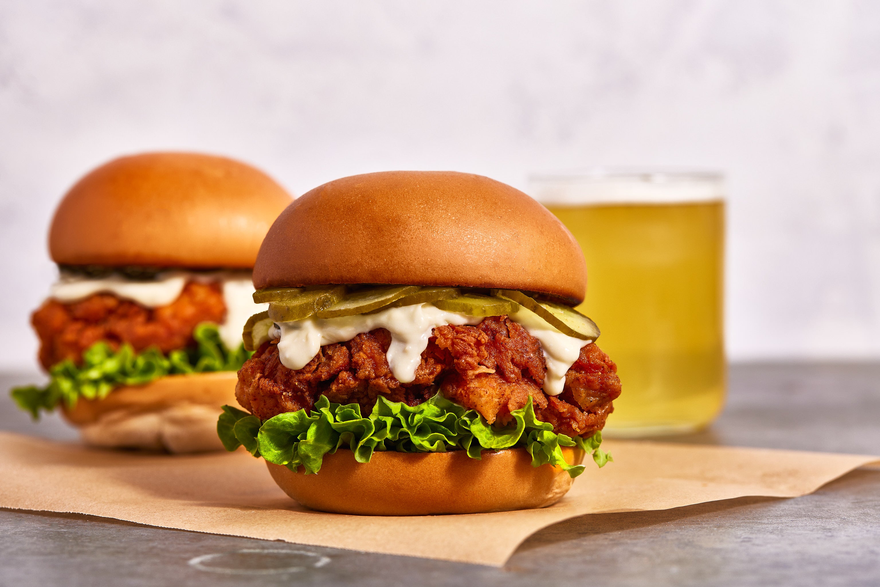 Two chicken burgers with lettuce and sauce on butter buns, served on a paper wrapper with a glass of beer in the background.