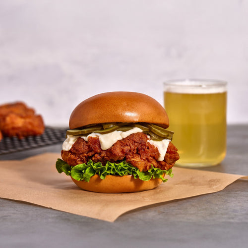 Burger with fried chicken, lettuce, and sauce on a bun, accompanied by a glass of beer and more fried chicken on a cooling rack.