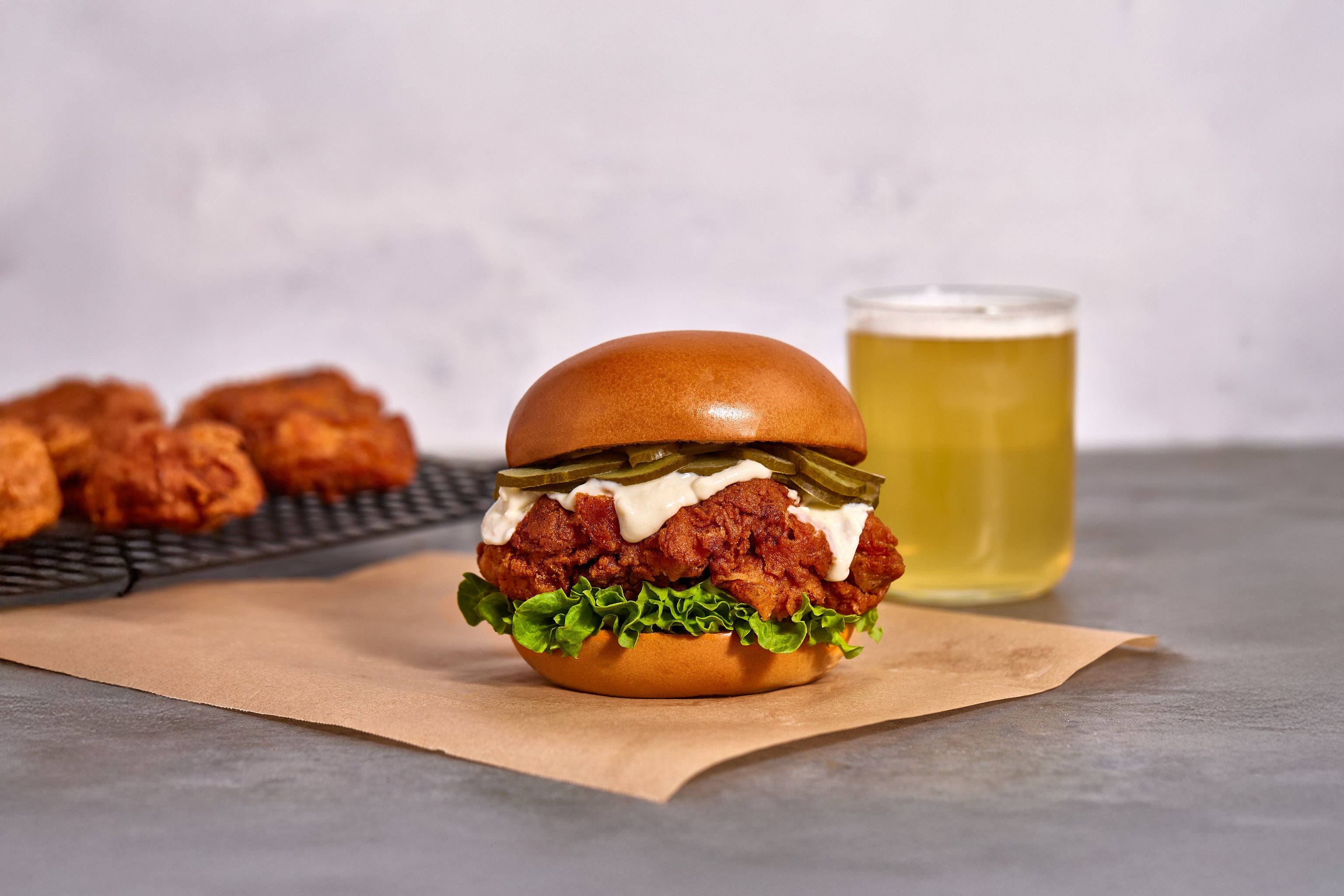 Burger with fried chicken, lettuce, and sauce on a bun, accompanied by a glass of beer and more fried chicken on a cooling rack.