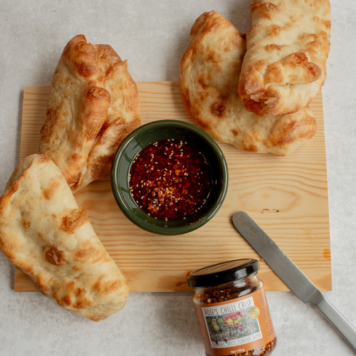 Three pieces of bread on a wooden board with a small bowl of red sauce and a jar of Chile paste with Acme knife.