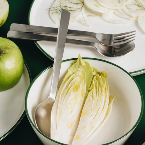 Acme utensils with halved endive lettuce on a white plate with green rim, surrounded by green apples and cut onions.