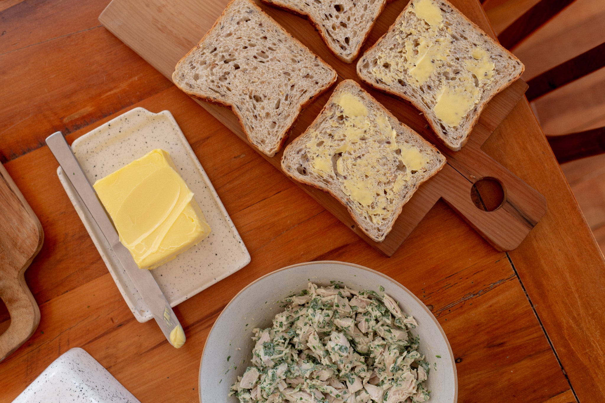 Butter on four slices of Volare Seeded Sandwich Loaf with a bowl of chicken mayo salad on a wooden table