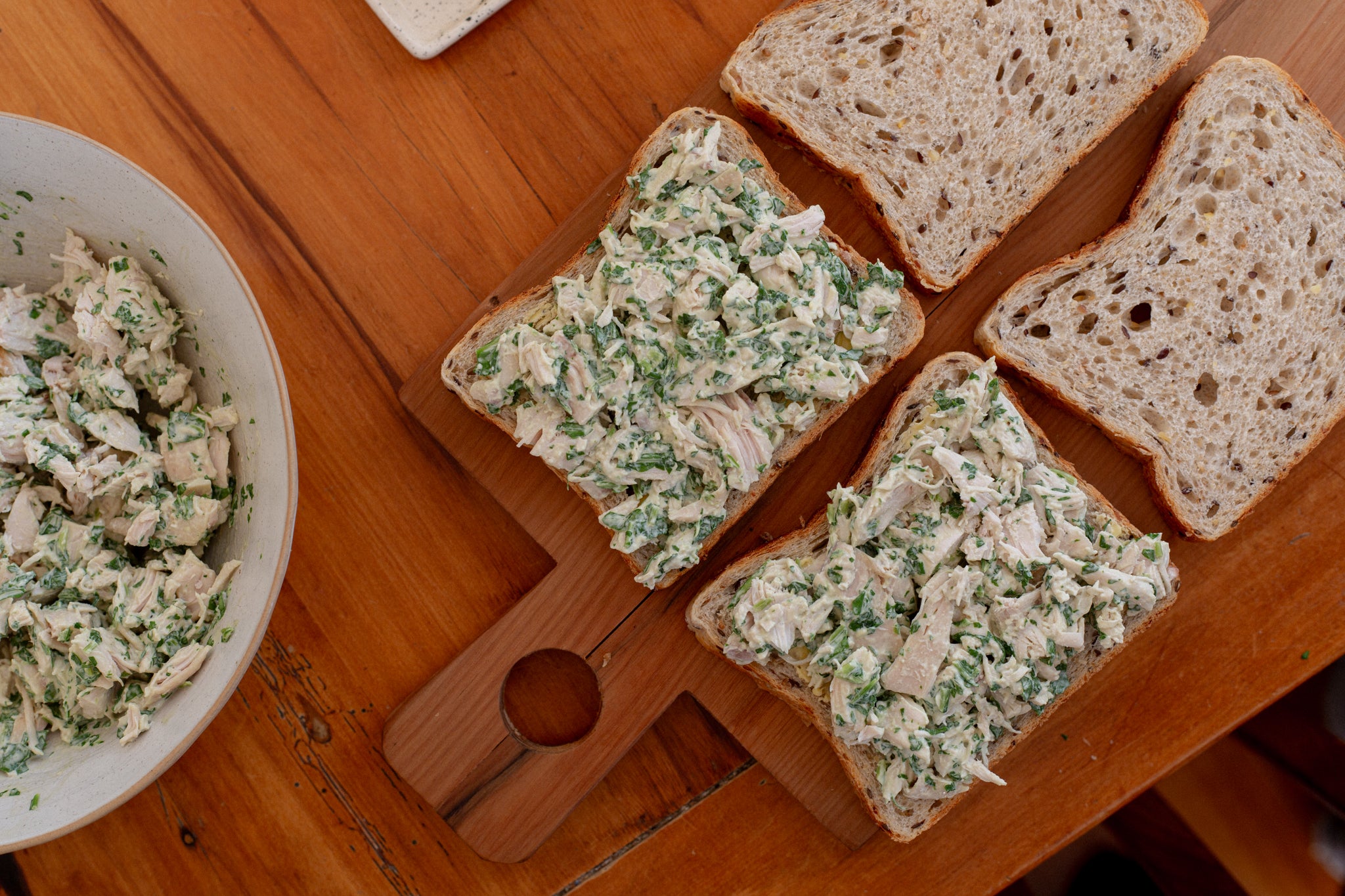 Four slices of Volare Seeded Sandwich Loaf with chicken salad on a wooden cutting board next to a bowl of chicken salad.
