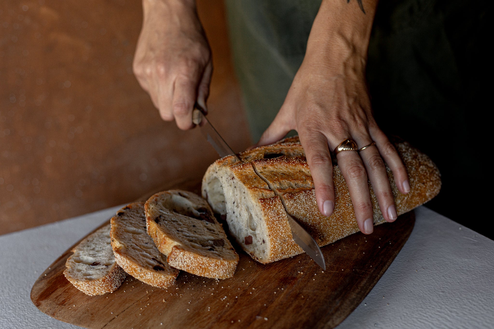 Volare Olive and Thyme Sourdough being sliced on a wooden cutting board with a bread knife.
