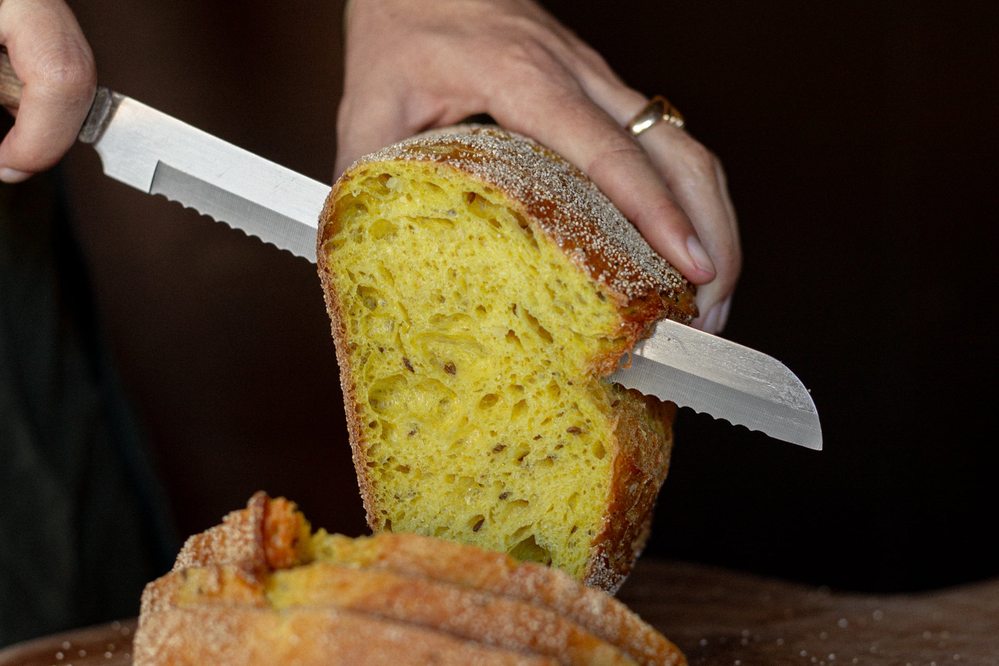 Volare Cumin, Onion & Turmeric Sourdough being cut with a bread knife on a dark background.