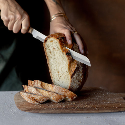 Person slicing Volare Roast Onion Sourdough on a wooden board with a knife.