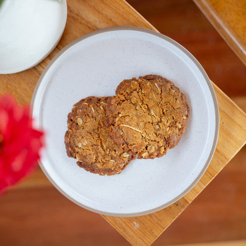 Two Volare ANZAC cookies on a white plate with flowers in the background