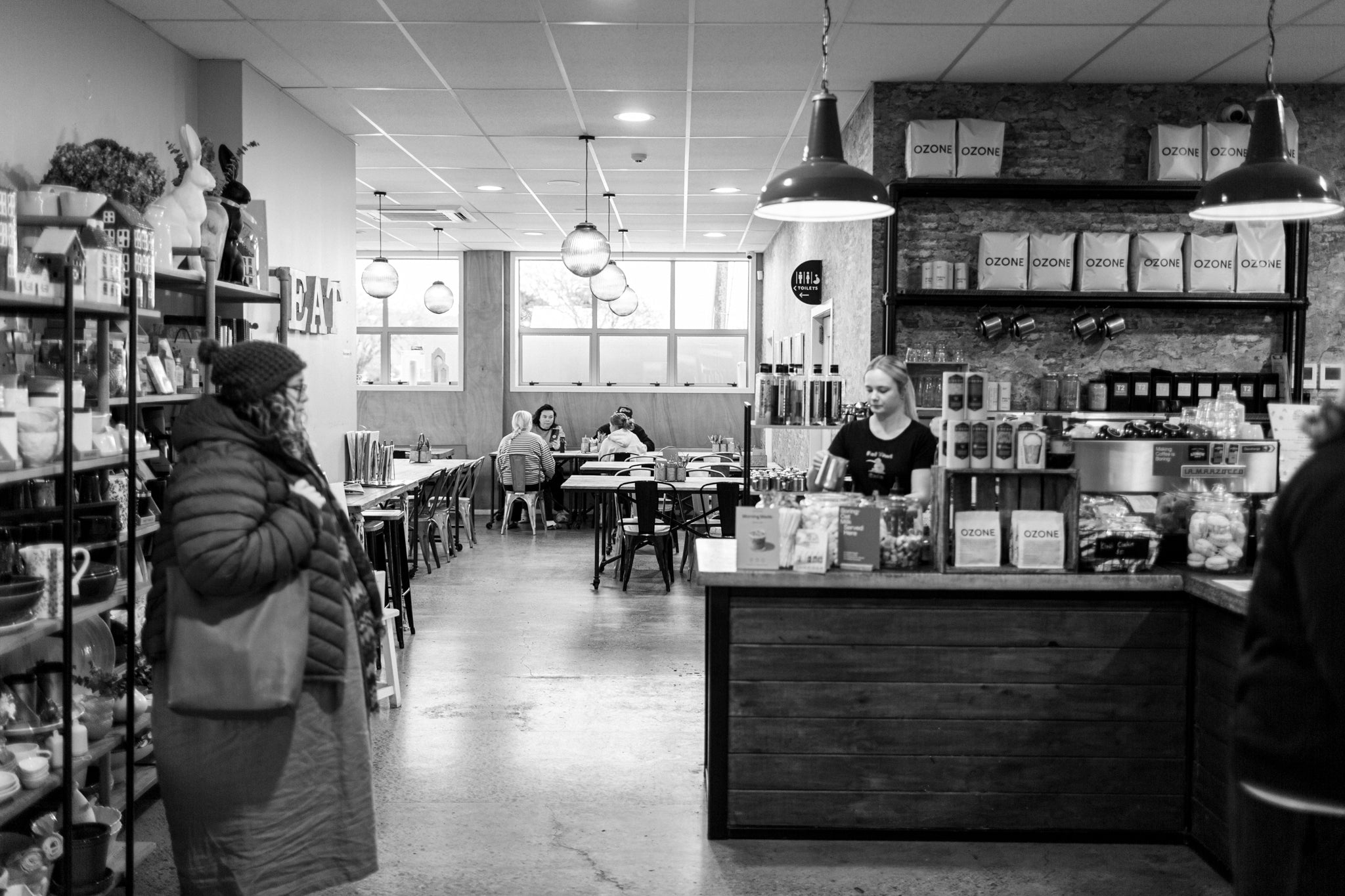A black and white photograph of a cozy Fat Kiwi Cafe interior with a wooden counter, hanging pendant lights, shelves, and customers and staff inside.