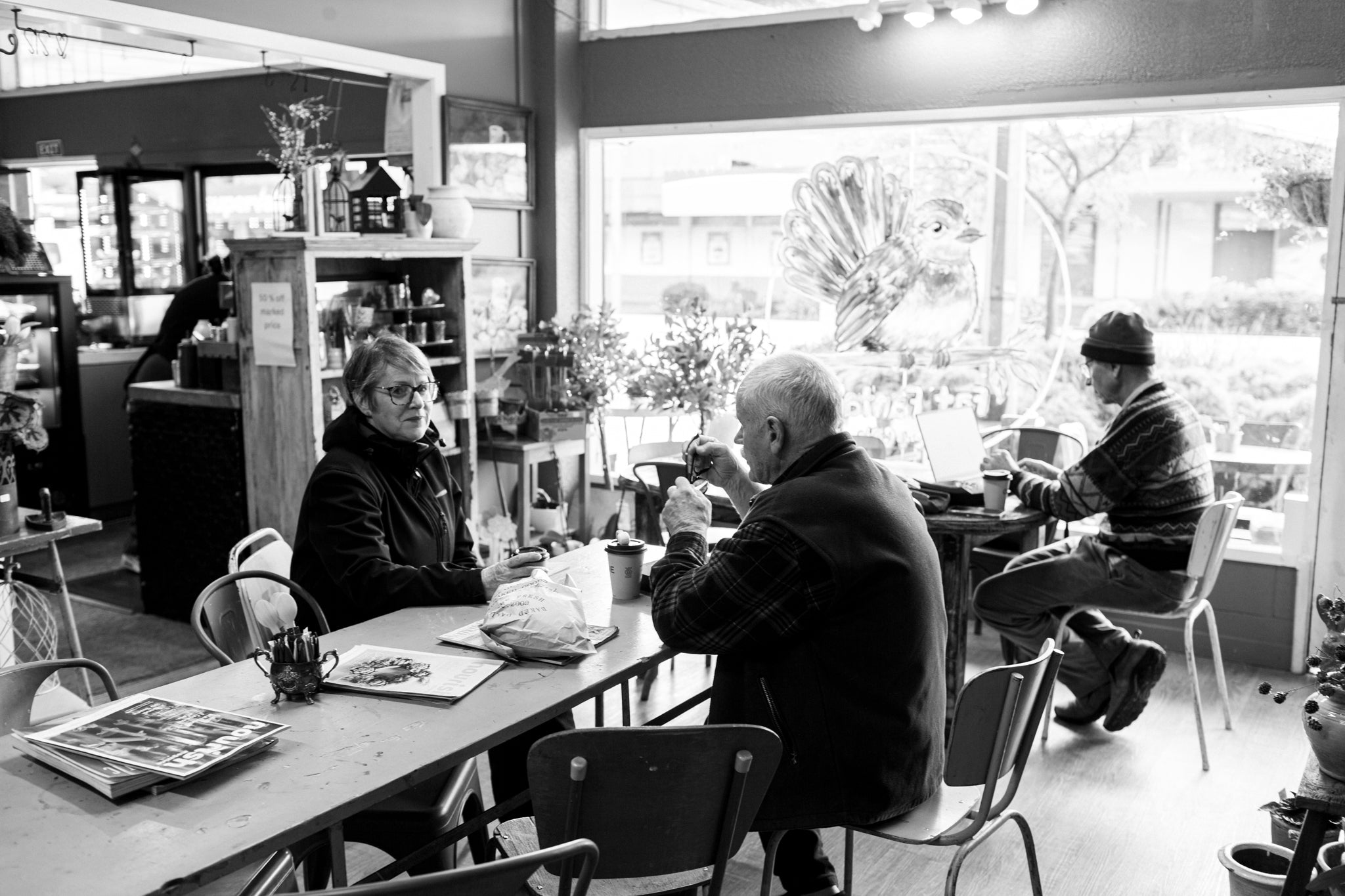 Black and white photograph of people sitting and working inside at Fat Fantail Gifts & Coffee, with large windows in the background.