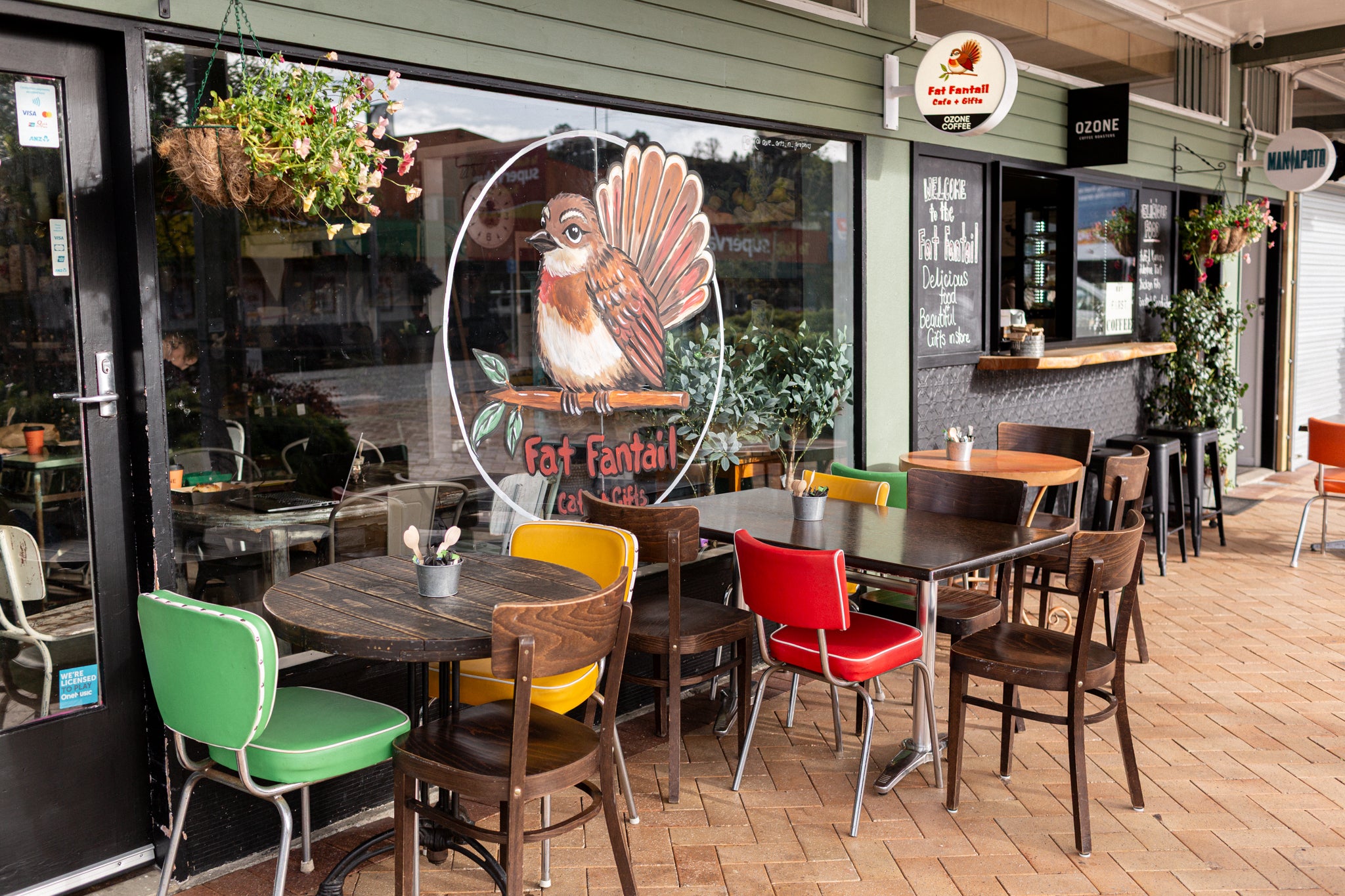Outdoor patio seating area of Fat Fantail Gifts & Coffee with wooden tables and colorful chairs in green, yellow, red, and blue, set against a storefront with a large bird mural on the window.