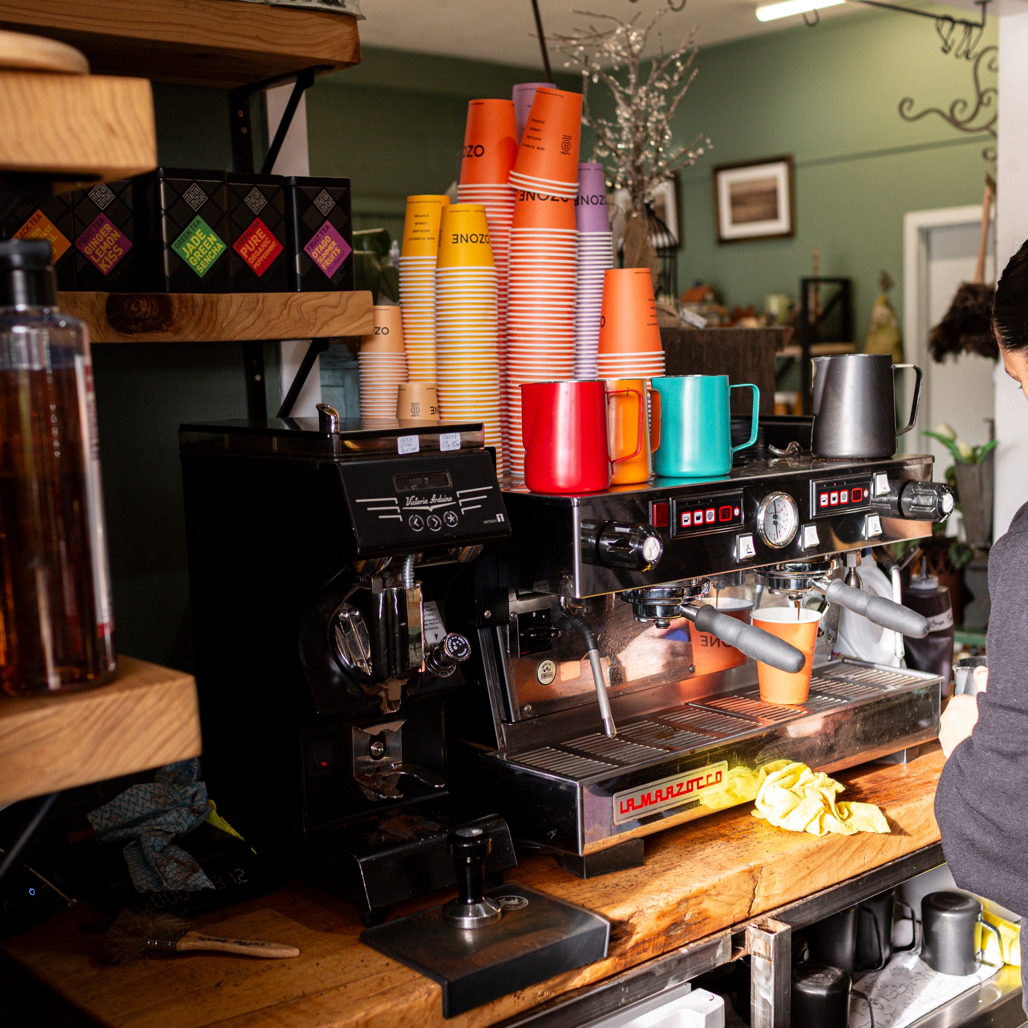 A black La Marche espresso machine on a wooden counter in a cafe setting, with stacks of colorful paper cups and mugs on top, shelves in the background, and a person partially visible on the right side.