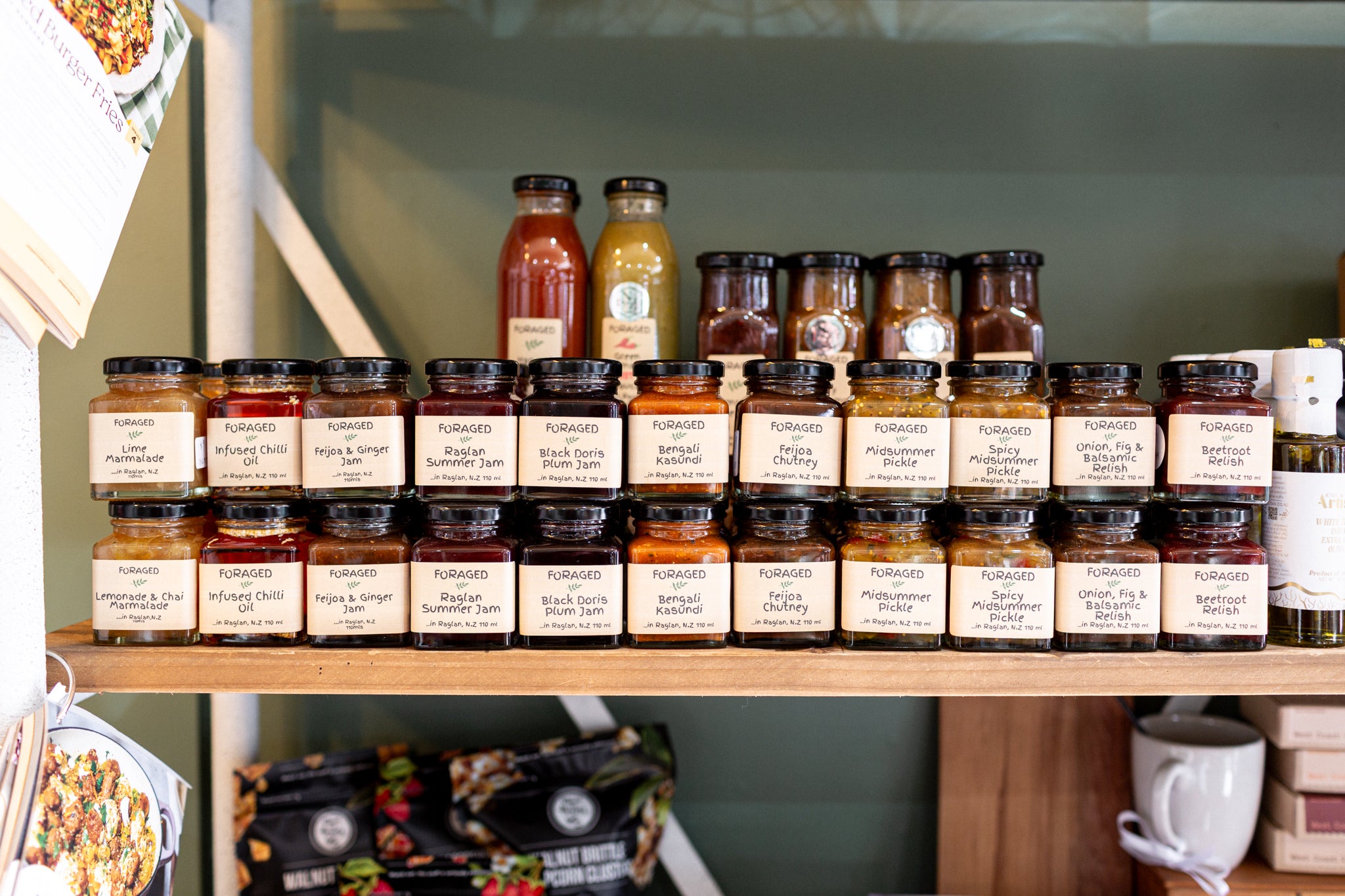 A shelf display of various gourmet spreads and sauces in glass jars with cream labels, alongside two tall bottles, set against a dark green background.