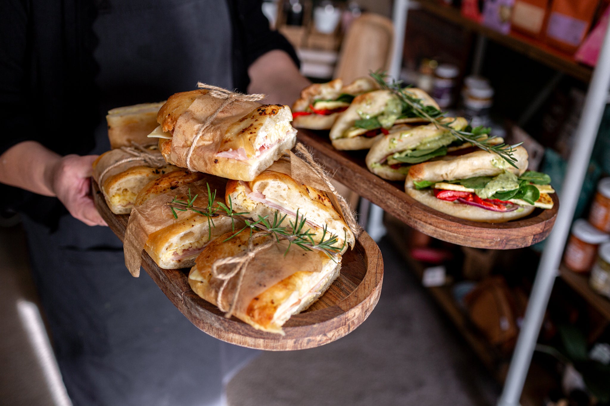 A person in a black shirt holds two wooden serving trays. One tray is filled with sliced bread rolls tied with twine and topped with rosemary.
