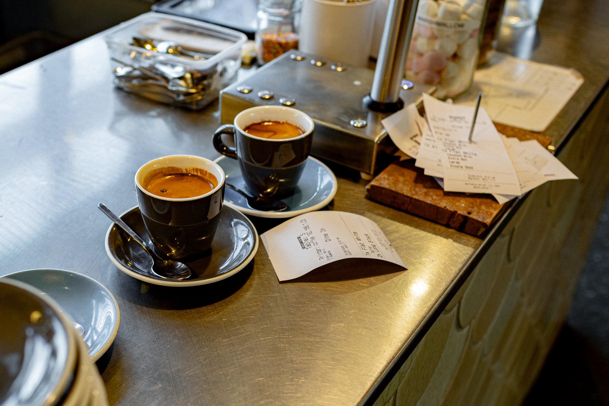 Two espresso cups on saucers on a stainless steel counter, with printed receipts.