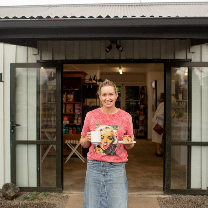 Owner of Clevedon Gourmet standing our front of the store holding a mug and plate with Hot Cross Bun
