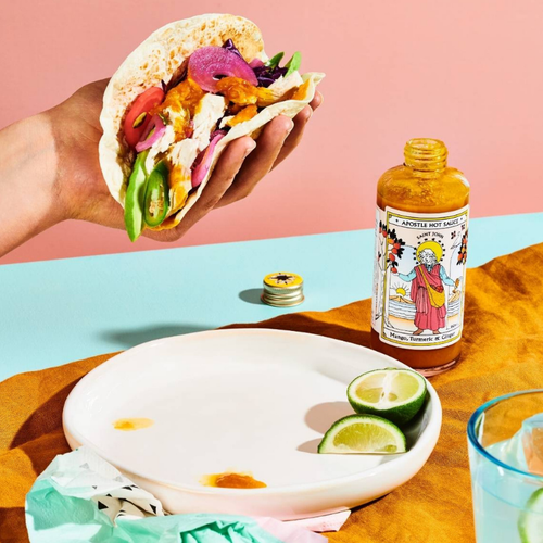 Hand holding a colorful taco with a bottle of hot sauce on a table.