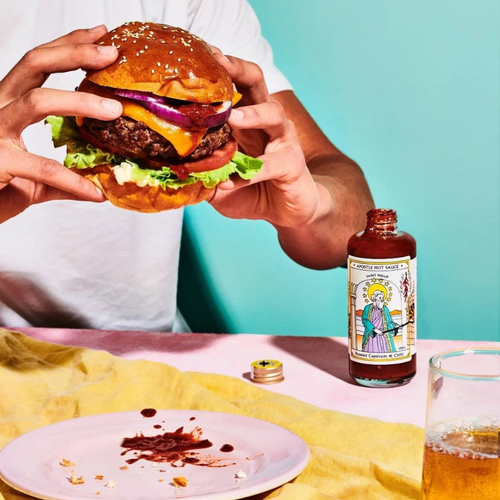 Person holding a burger with a bottle of hot sauce on a table