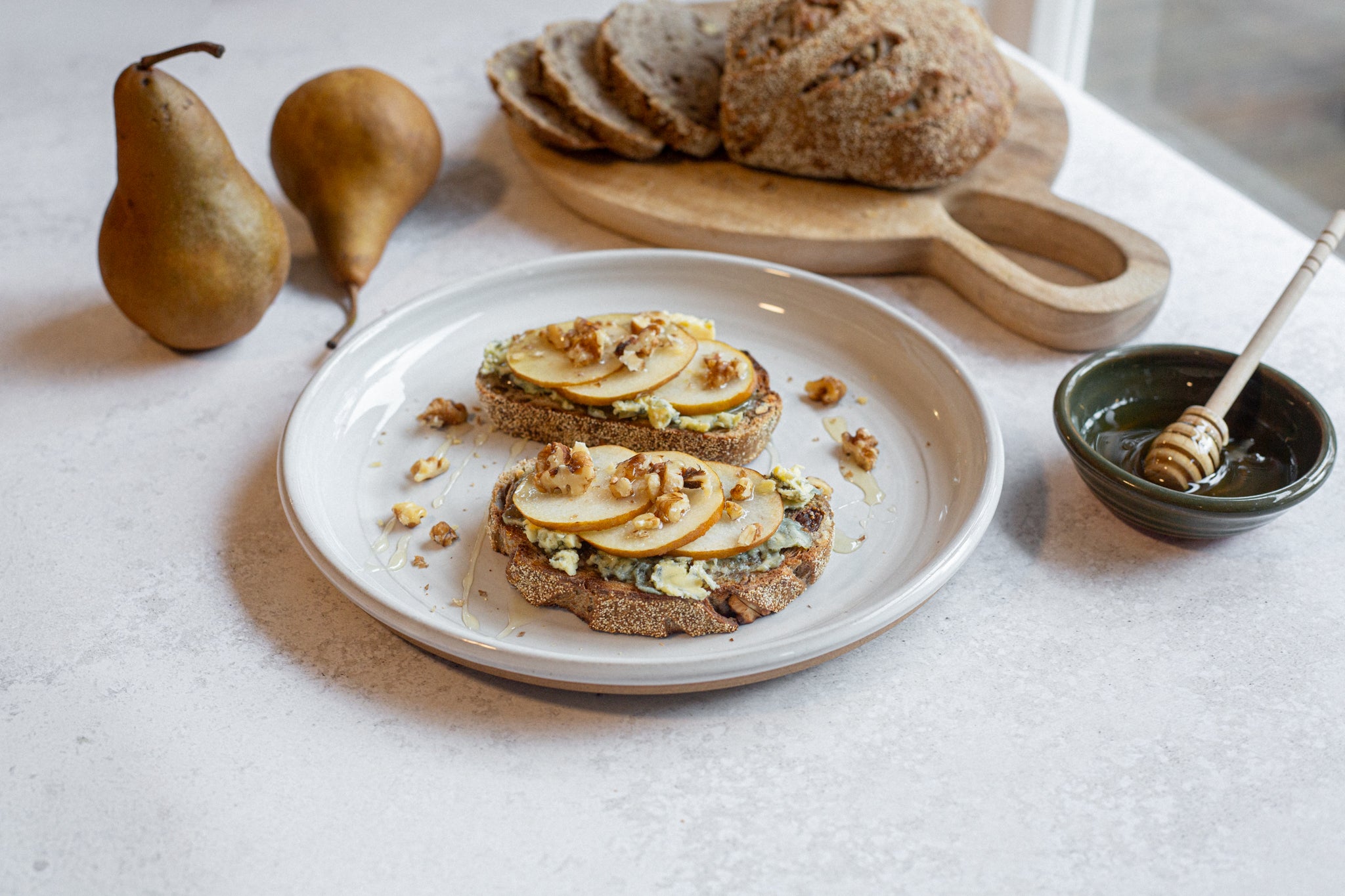 Two slices of bread with pear and walnut toppings on a white plate, accompanied by pears and a bowl of honey on a light surface.