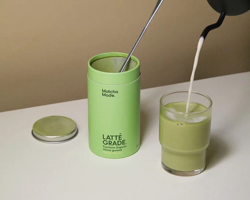 Person pouring milk into a glass of matcha green tea next to a container labeled 