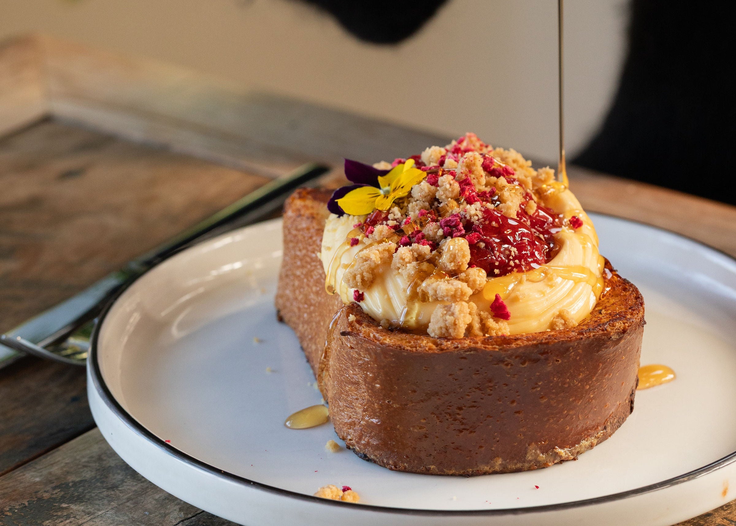 A close-up of a Punnets french toast on a white plate with a thin black rim, featuring a thick slice of brioche, cream-colored topping, and red berry crumble, with a hand pouring dark syrup from a small metal pitcher.
