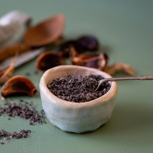Small ceramic bowl filled with Opito Bay Black Garlic Sea Salt on a green surface with cinnamon sticks.