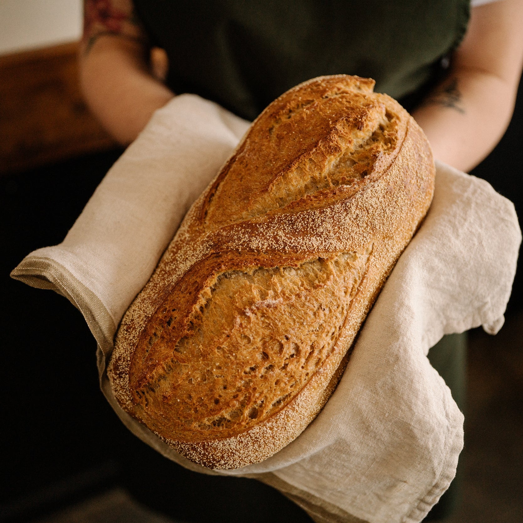 A person in a dark green apron holds a freshly baked sourdough bread loaf wrapped in a white cloth towel in a warm, dimly lit kitchen setting.