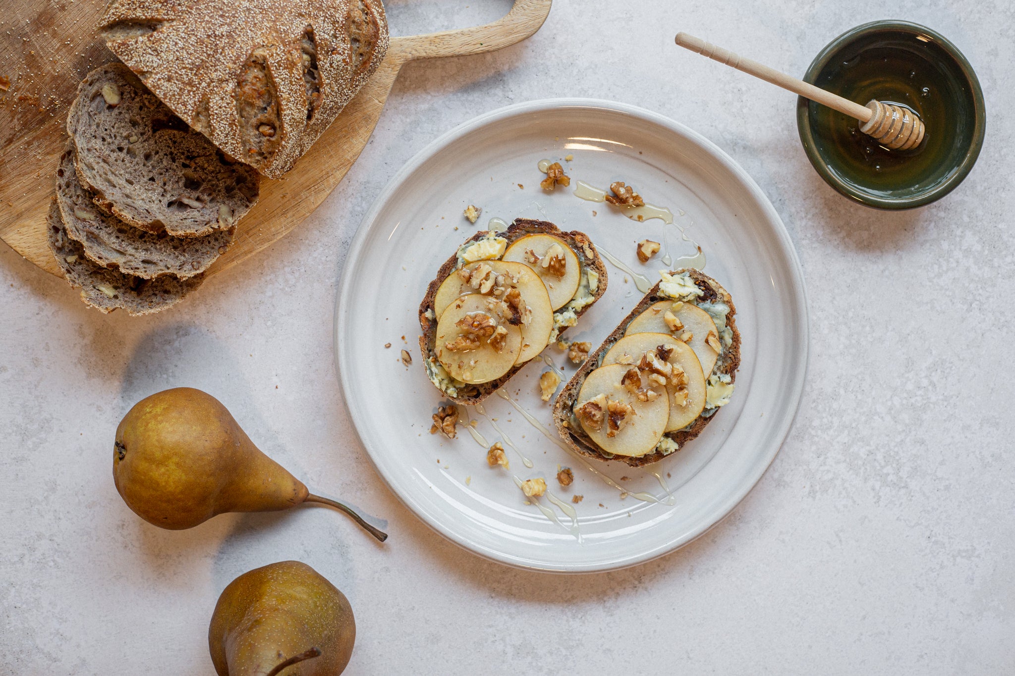 Plate of bread with pear slices and nuts on a light surface with pears and a honey dipper.